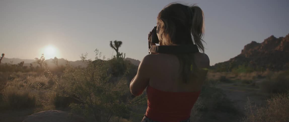 A woman holds a camera to her face taking a picture of a low sun in a rural area