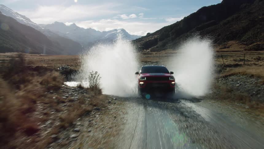 Car drives along dirt road past fenced pasture scrubby field and through stream with mountains in background