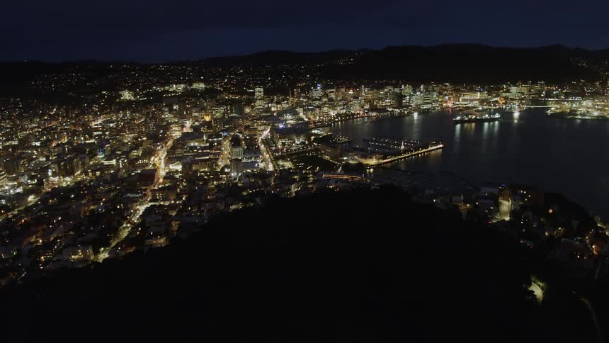 The silhouette of a hill reveals a waterfront city with marina and lights shimmering at night