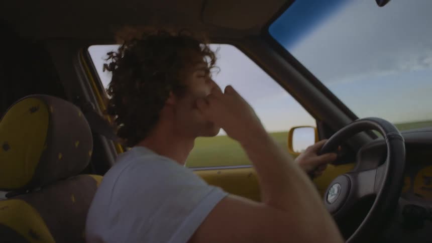 A man in a vehicle with a steering wheel with a grassy field the sky and clouds in the background