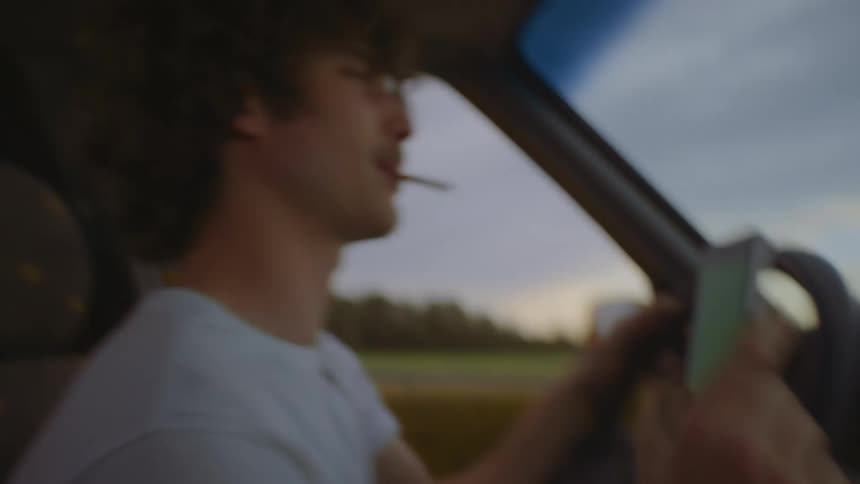 A smoking man dances and checks his phone while driving a car through the countryside during the day