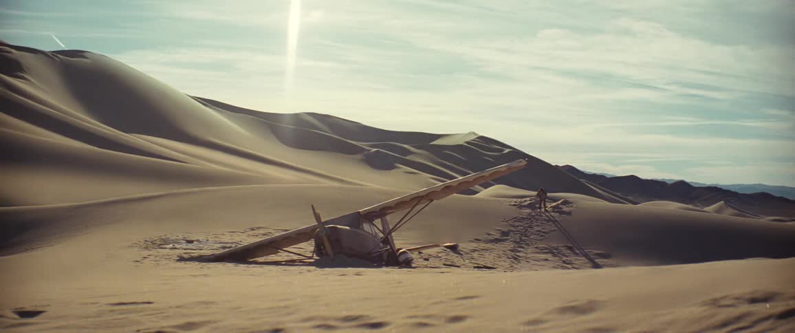 Man sits down next to a damaged airplane in the desert as another person walks towards him on the sand