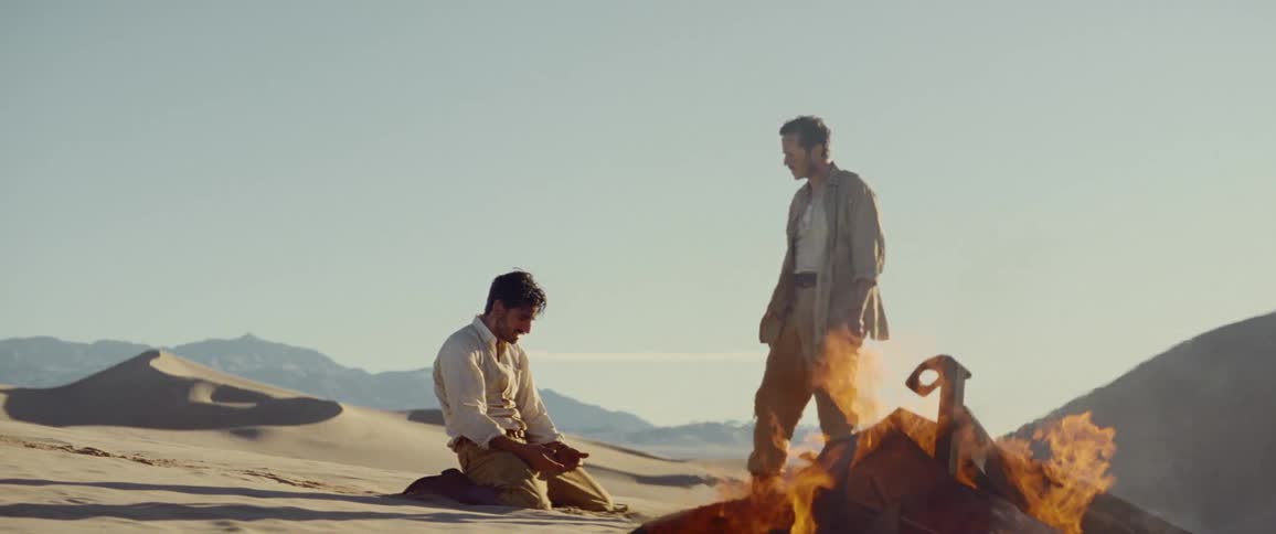 A man kneels in the desert next to burning wreckage as another man stands over him
