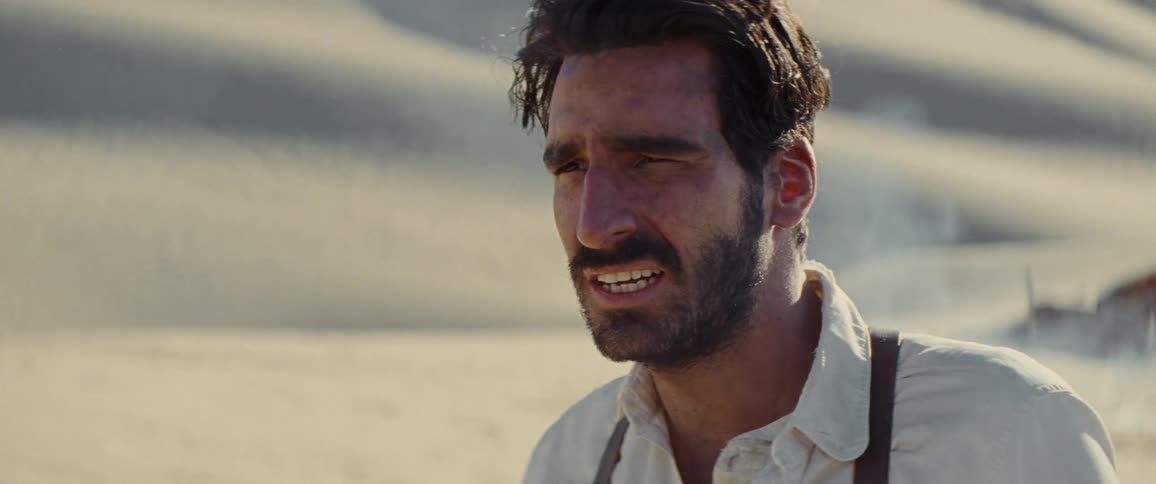 Bearded man wearing a wristwatch shields his eyes from the sun in the desert as smoke billows behind him