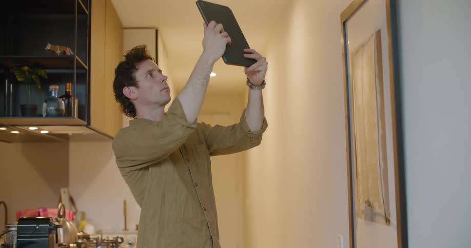 Man stands in a kitchen and holds up a tablet and looks at its screen