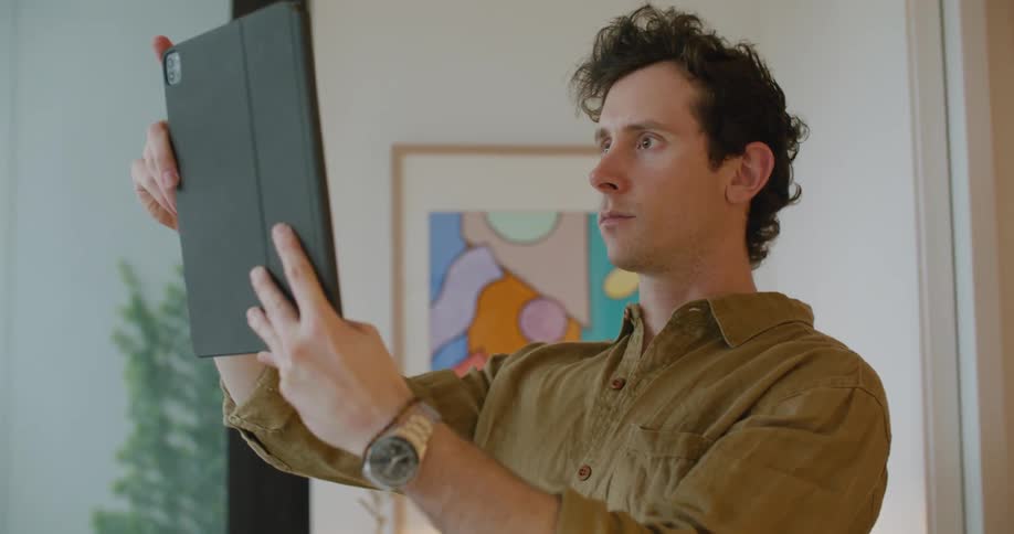 A man in a room using a tablet with a wall and picture frame in the background