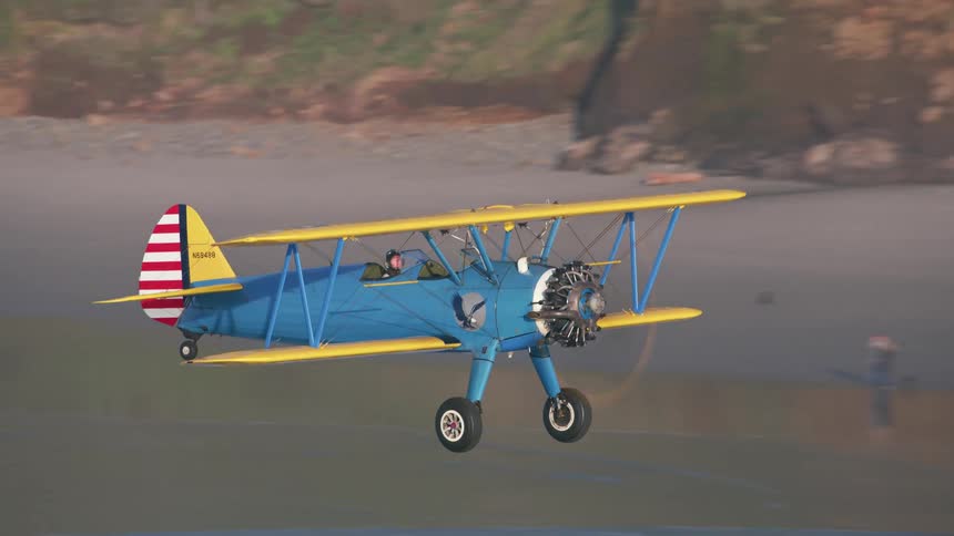 A person with a headset flying a plane over a beach with vehicles a road and cliffs in the background