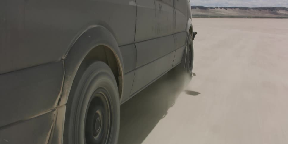 A vehicle speeds off road across a barren flatland during the day