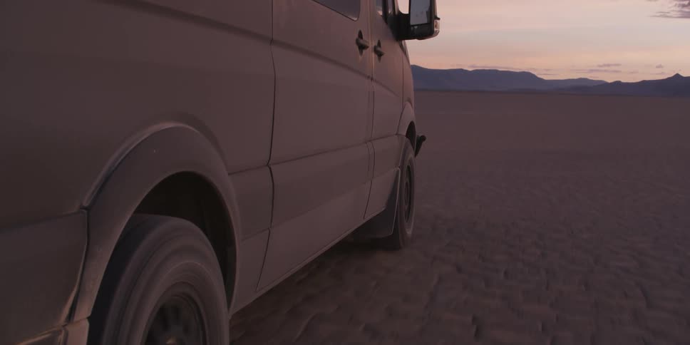 A dusty and dirty van drives a long a wide open desert landscape with cracked soil