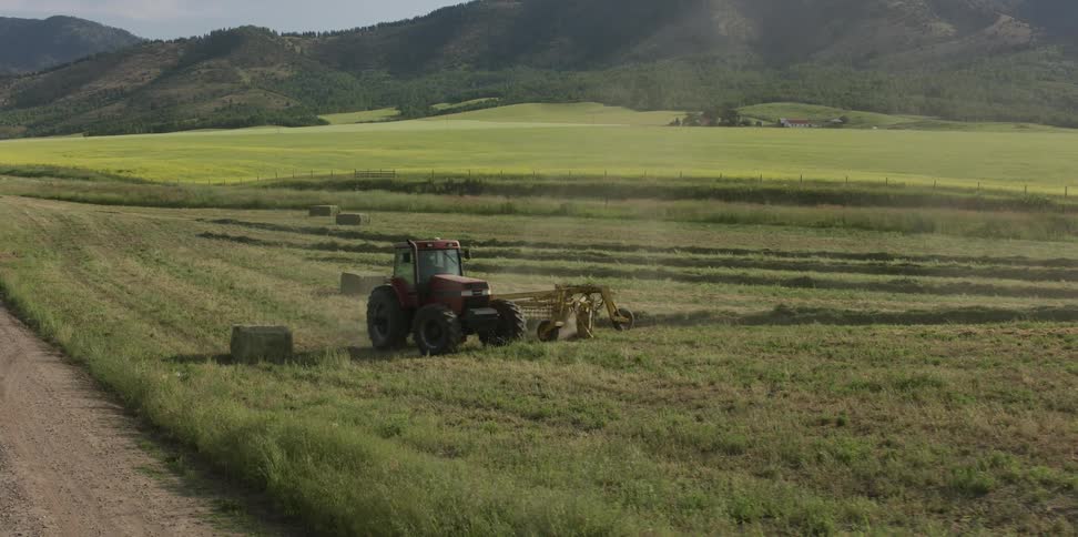 Truck drives past in background between tractor towing mowing apparatusand forested mountains