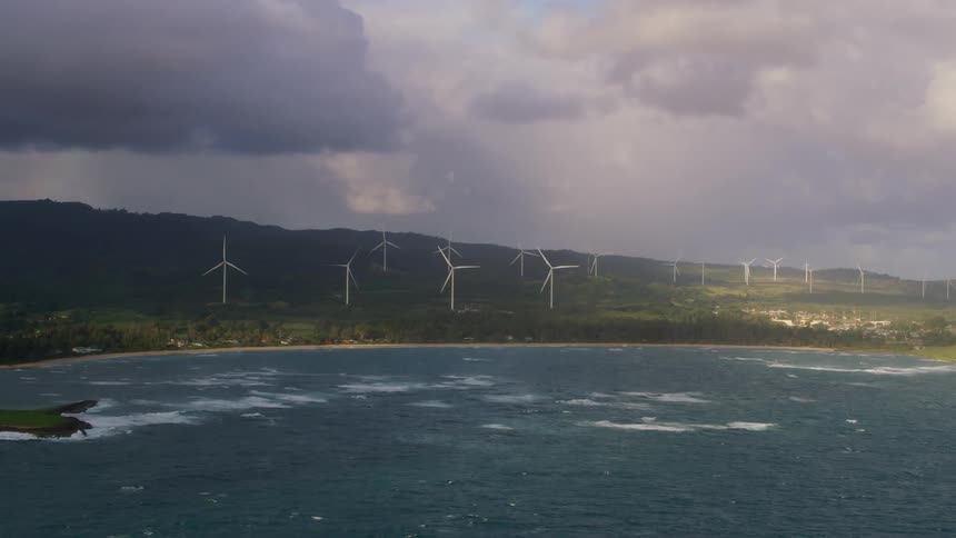 Waves roll into shore as turbines on a wind farm are spinning under a cloudy sky