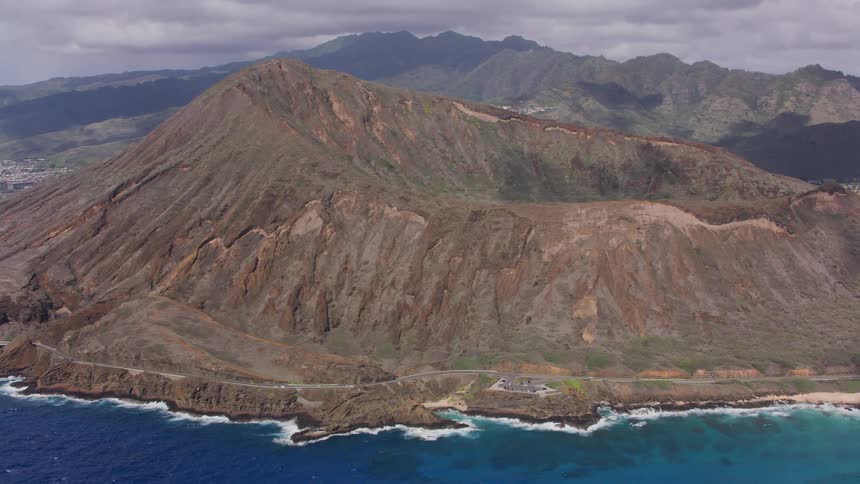 Waves splash on shore with a cratered mountain and cars traveling on a coastal road