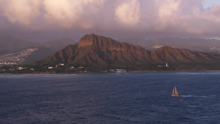 A sailboat in the ocean with land buildings the sky and clouds in the background