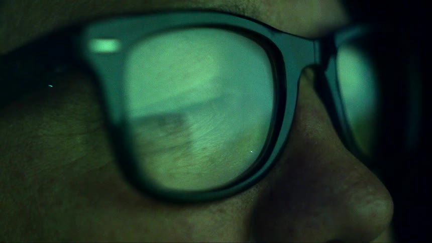 Close up of red and green lights reflecting over the glasses of a man inside a dark room