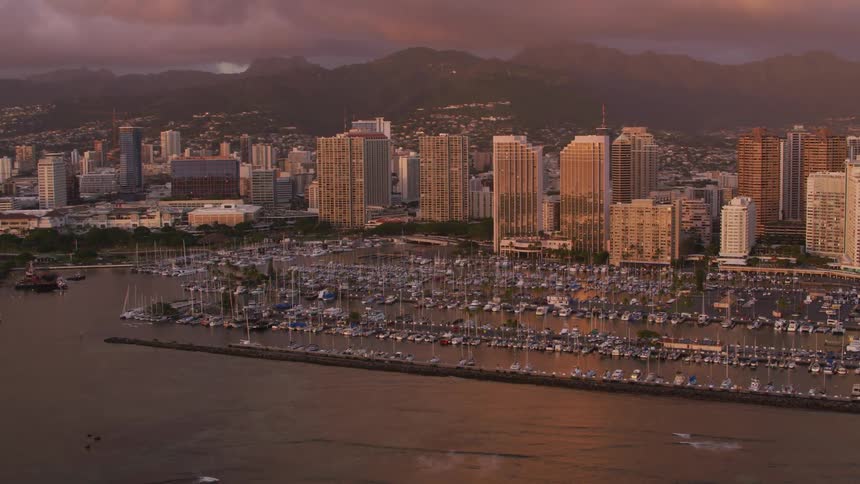 A coastal city with a marina in the foreground with a mountain range the sky and clouds in the background