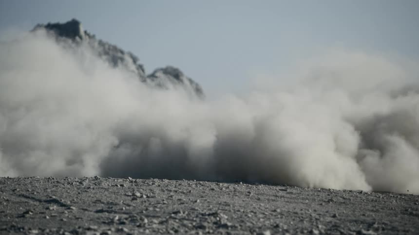 Dust cloud flowing across dirt and gravel partly obscures mountain in background