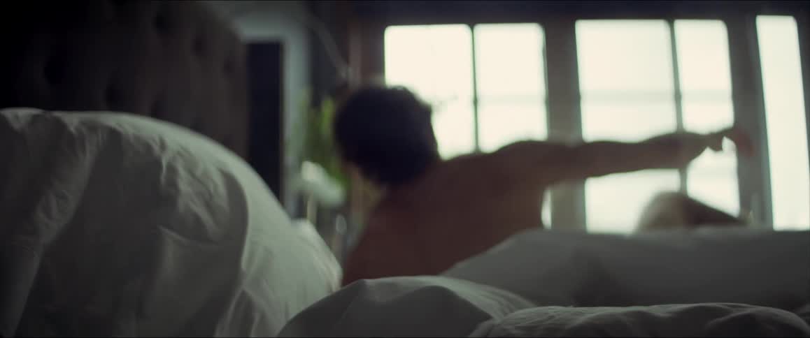 A man rolls over in bed and turns his alarm off and gets out of bed and stands and stretches his arms up