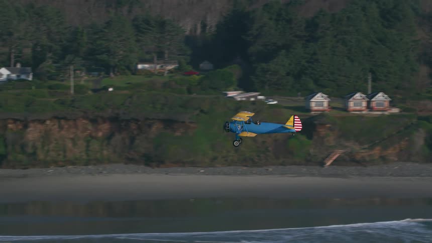 An airplane flying over a beach with waves with houses and trees in the background