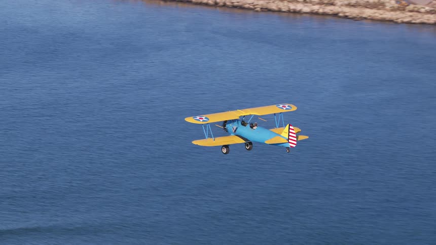 Colorfully painted biplane flies over ocean paralleling shore passing forest and dirt road
