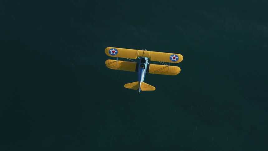 A brightly painted biplane flies at low altitude above the ocean water