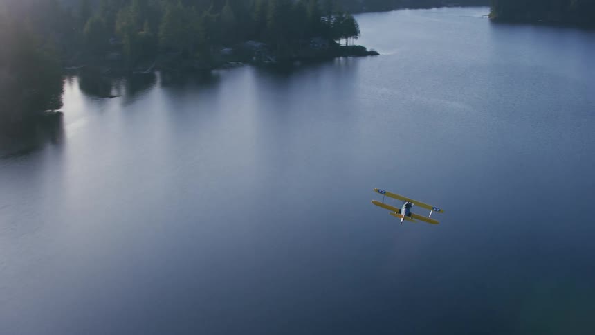 A biplane flies over a river through a forest during the day