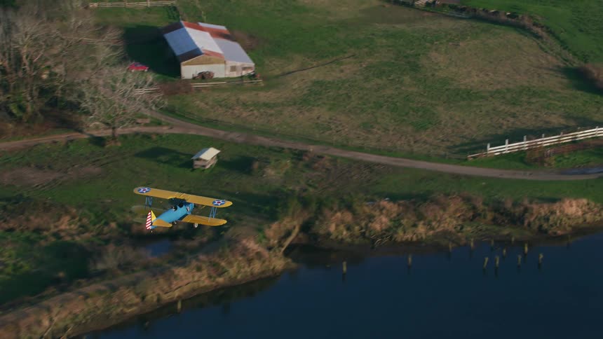 Yellow and blue biplane flies over river pastures and farm