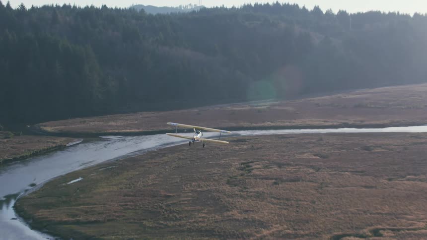 An airplane flies over a creek in an open rural area