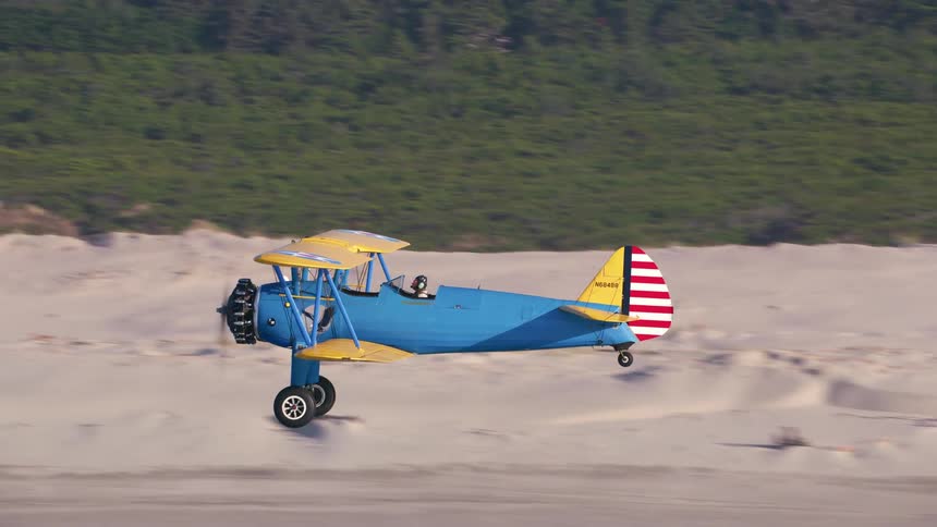 Person pilots blue and yellow biplane low over ocean near shore beach with forest in background