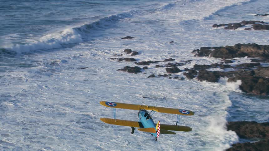An aerial view of a biplane flying along a rocky coastline with waves crashing onto the rocks