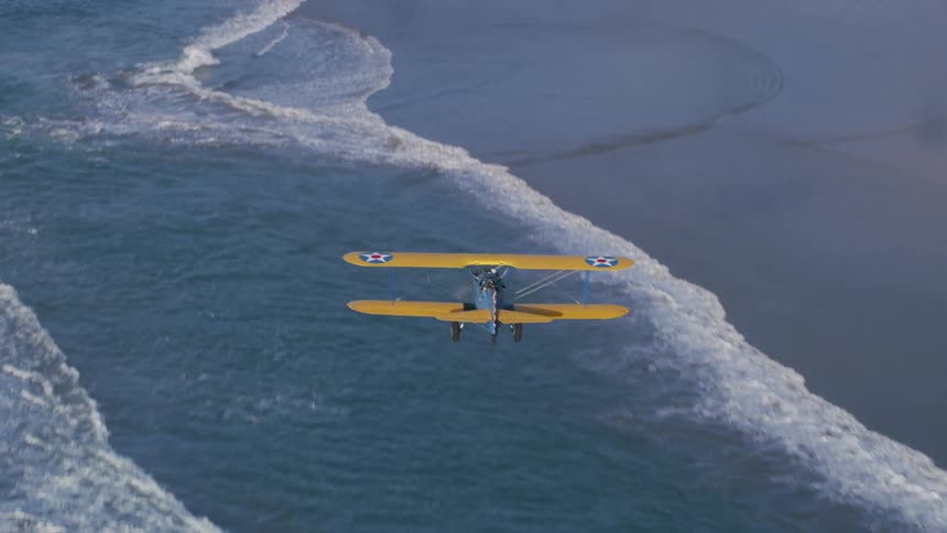 Yellow and blue biplane flies over ocean waves and surf then over slightly reflective wet sand of beach