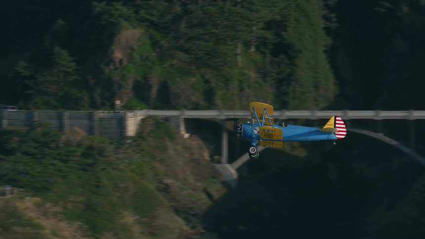 A vintage warbirds biplane flies over hills bridge and road