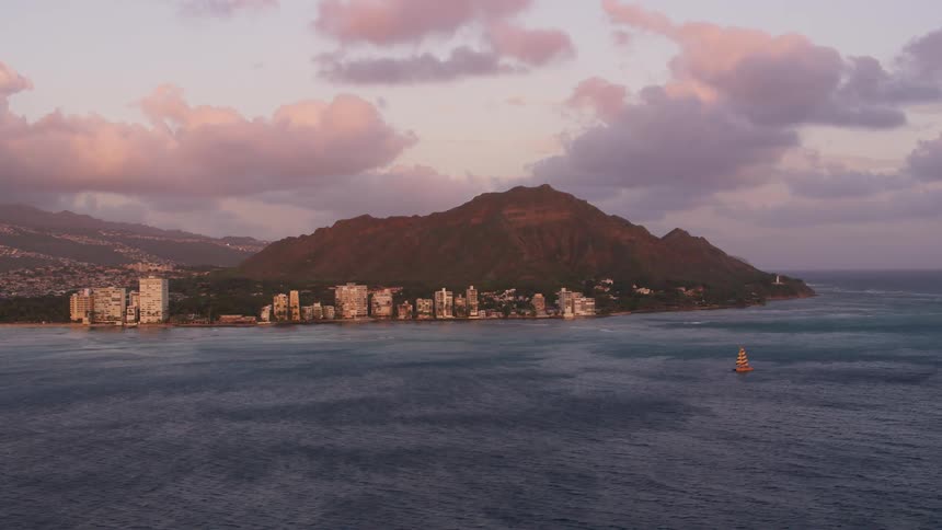 A sailboat in the ocean with land buildings the sky and clouds in the background