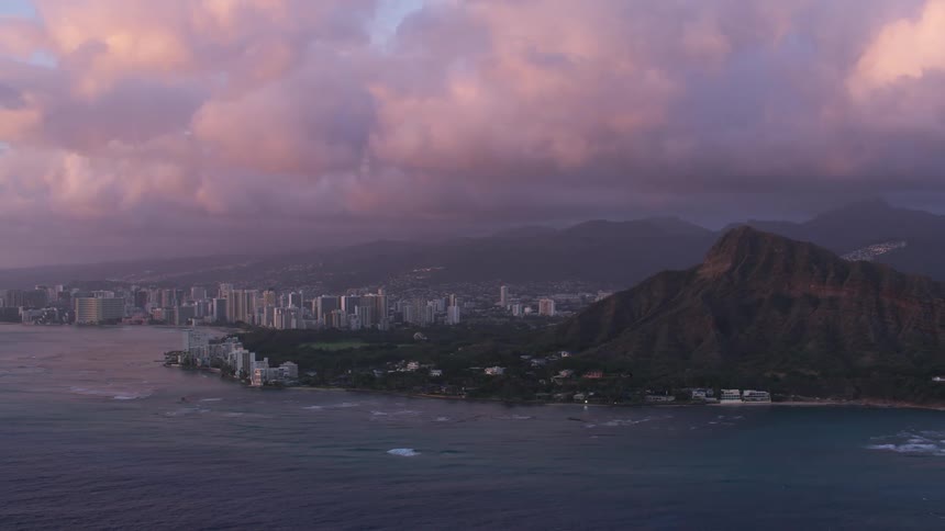 An aerial view of large coastal city with mountains and puffy clouds at dusk