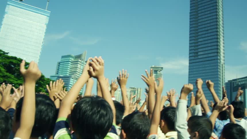 A large crowd of children wave their hands in the sky by the city