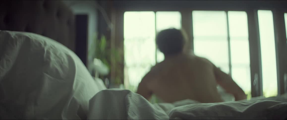 A man gets out of bed wearing his boxers and stretches in front of large windows