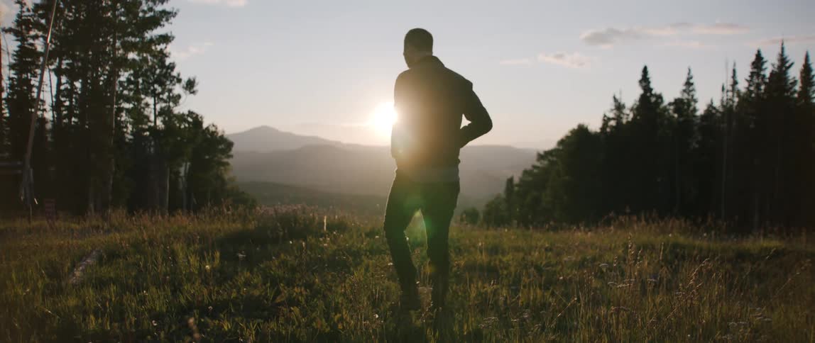 A silhouette of a man walking on a grassy field with trees the sky and clouds in the background