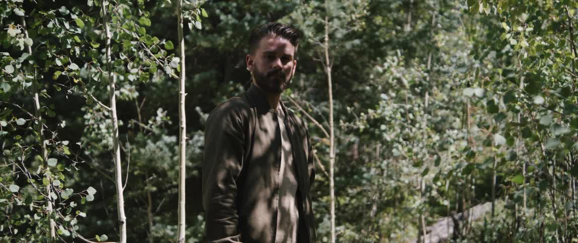 Bearded man wearing jacket stands among young trees looking