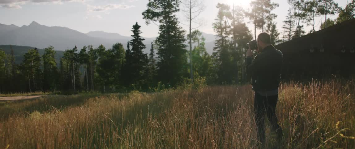 Young man filming the grass on the field near the forest on the mountains at sunset