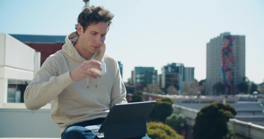A man sits on a balcony ledge with a laptop computer and takes a drink from a small mug