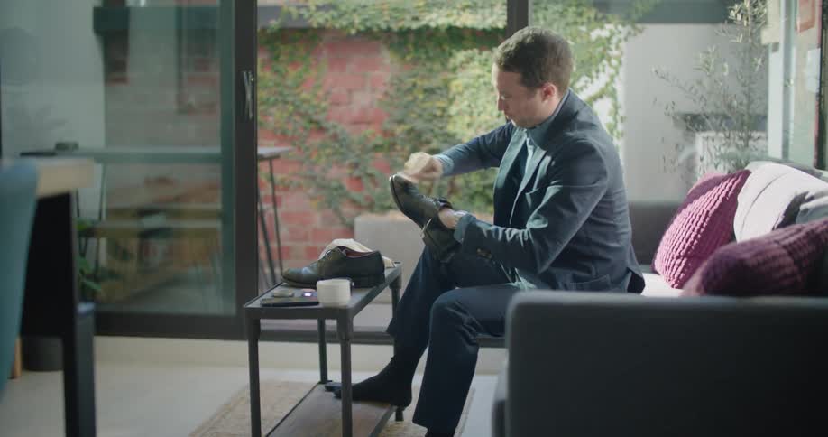 A man sits on a couch at home and polishes his shoes