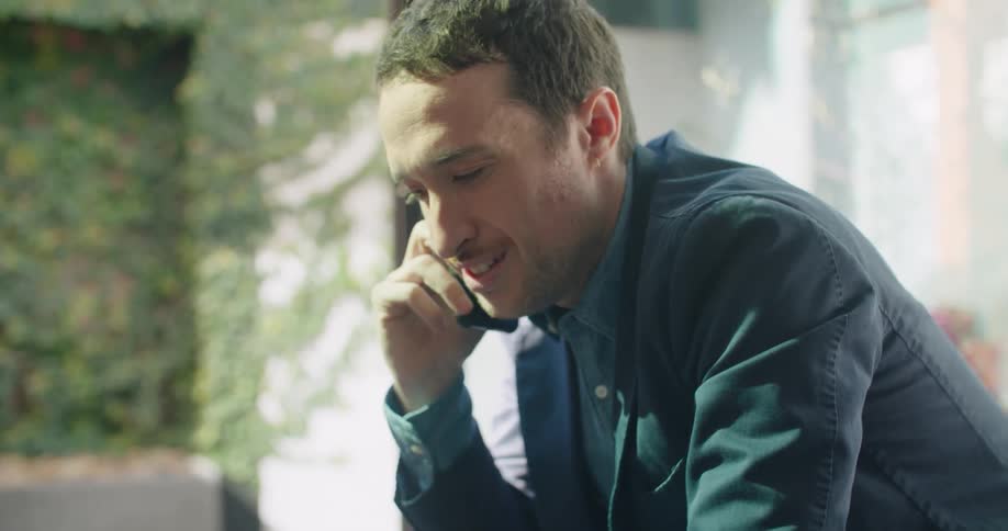 A man talking on a smart phone and using a laptop on a table with a wall in the background