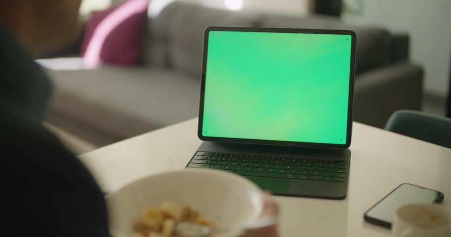A man is sitting at a table inside a home while having breakfast cereal and looking at a laptop