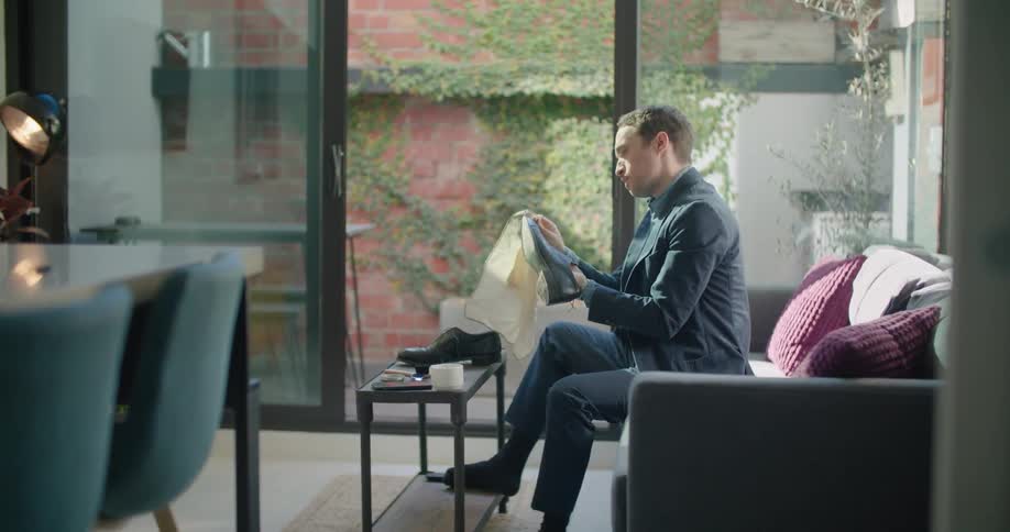 A man in a suit shines his shoes while sitting on the couch