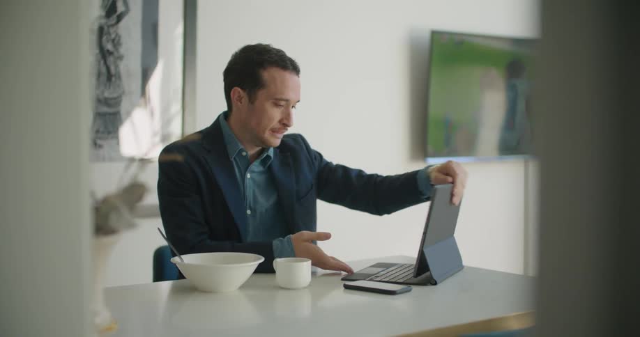 A man sitting at a table drinks from a coffee cup while looking at his laptop before checking his watch and carrying his laptop away