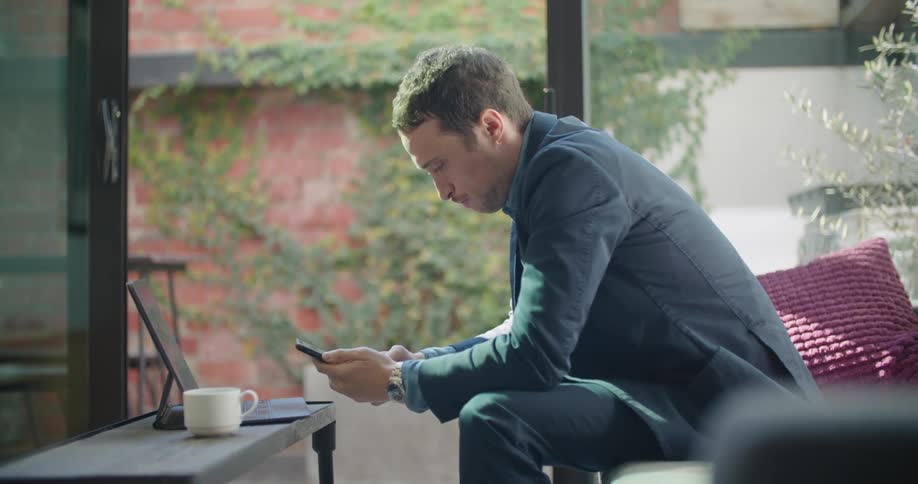 A man sits working on a laptop and then picks up a cell phone to make a call