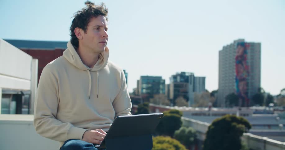 A man is sitting on a balcony overlooking a city and typing on his laptop