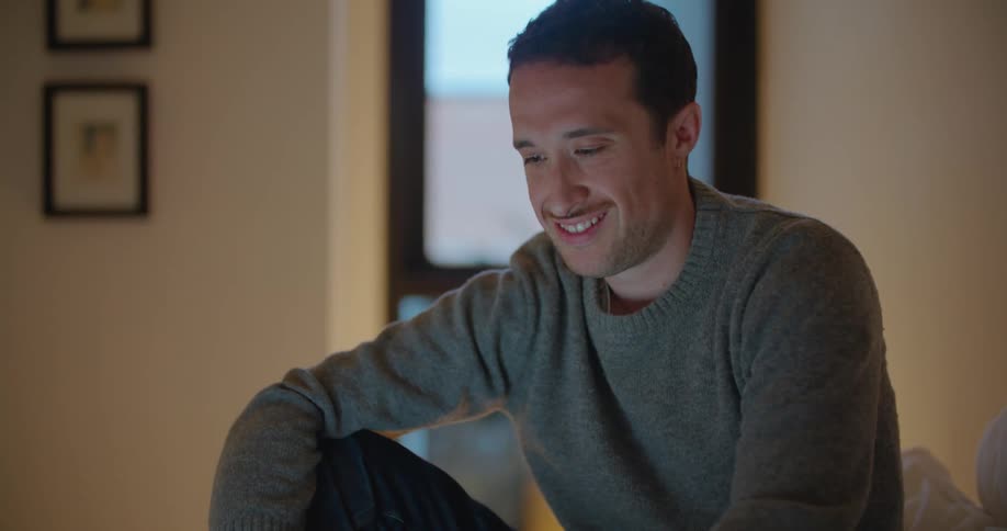 A man in a room sitting on a bed using a laptop with a wall and window in the background