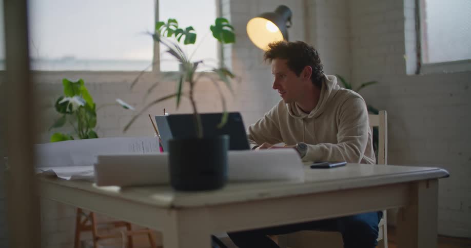 A man in a room sitting at a table using a laptop with a wall and window in the background