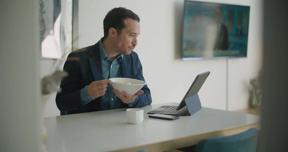 Man sits at table with bowl taking a few bites before concentration on tablet