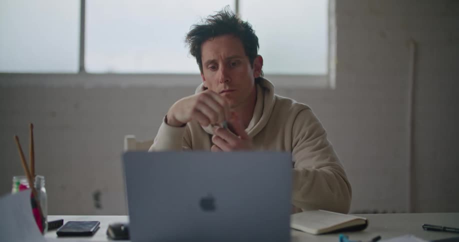 A man scratches his head and clicks a pen while working on his laptop